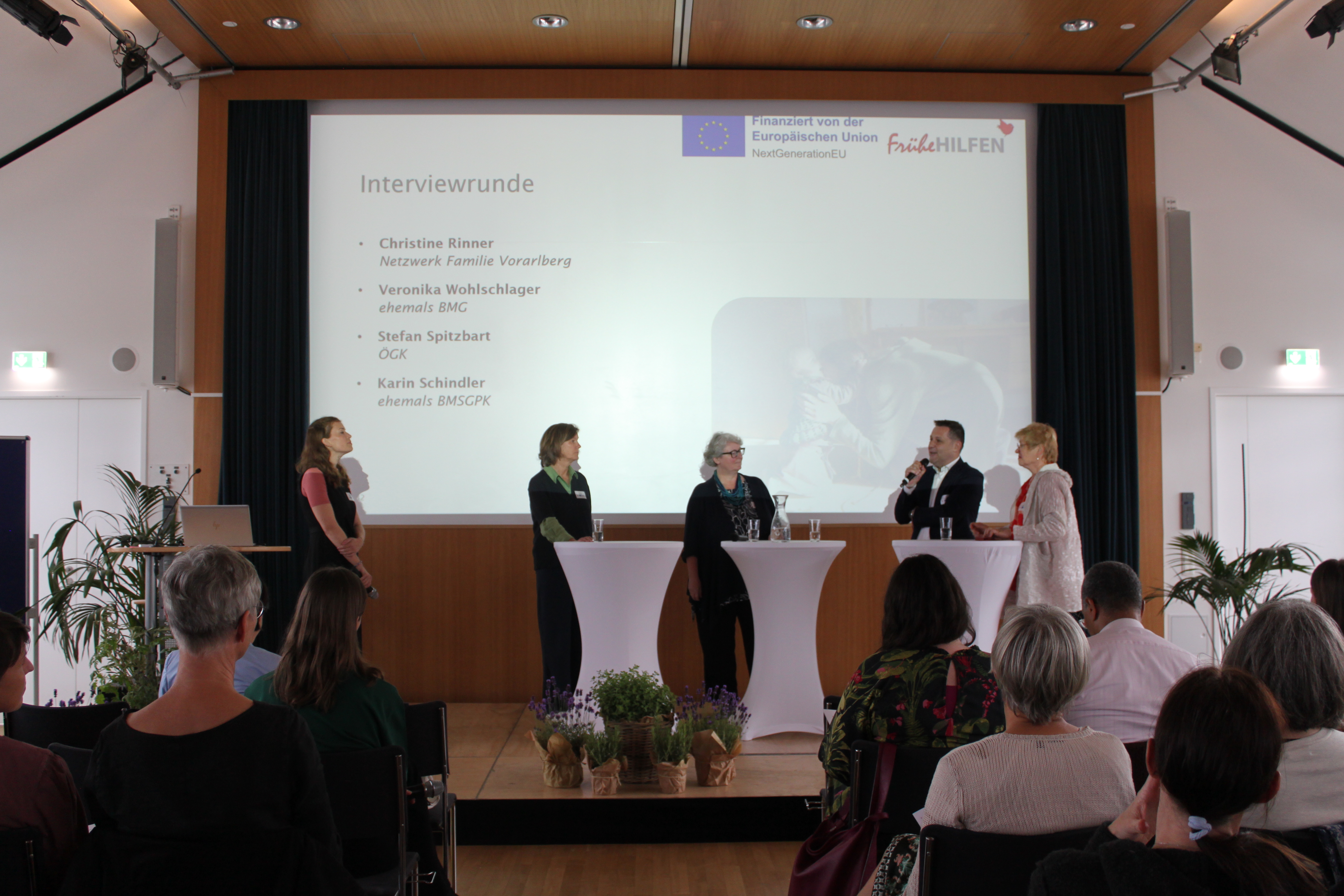 Podiumsgespräch bei der Abschlussveranstaltung zum österreichweiten Ausbau der Frühen Hilfen. Mehrere Personen stehen auf einer Bühne vor einer Präsentationsfolie zur Interviewrunde, während das Publikum die Diskussion verfolgt.