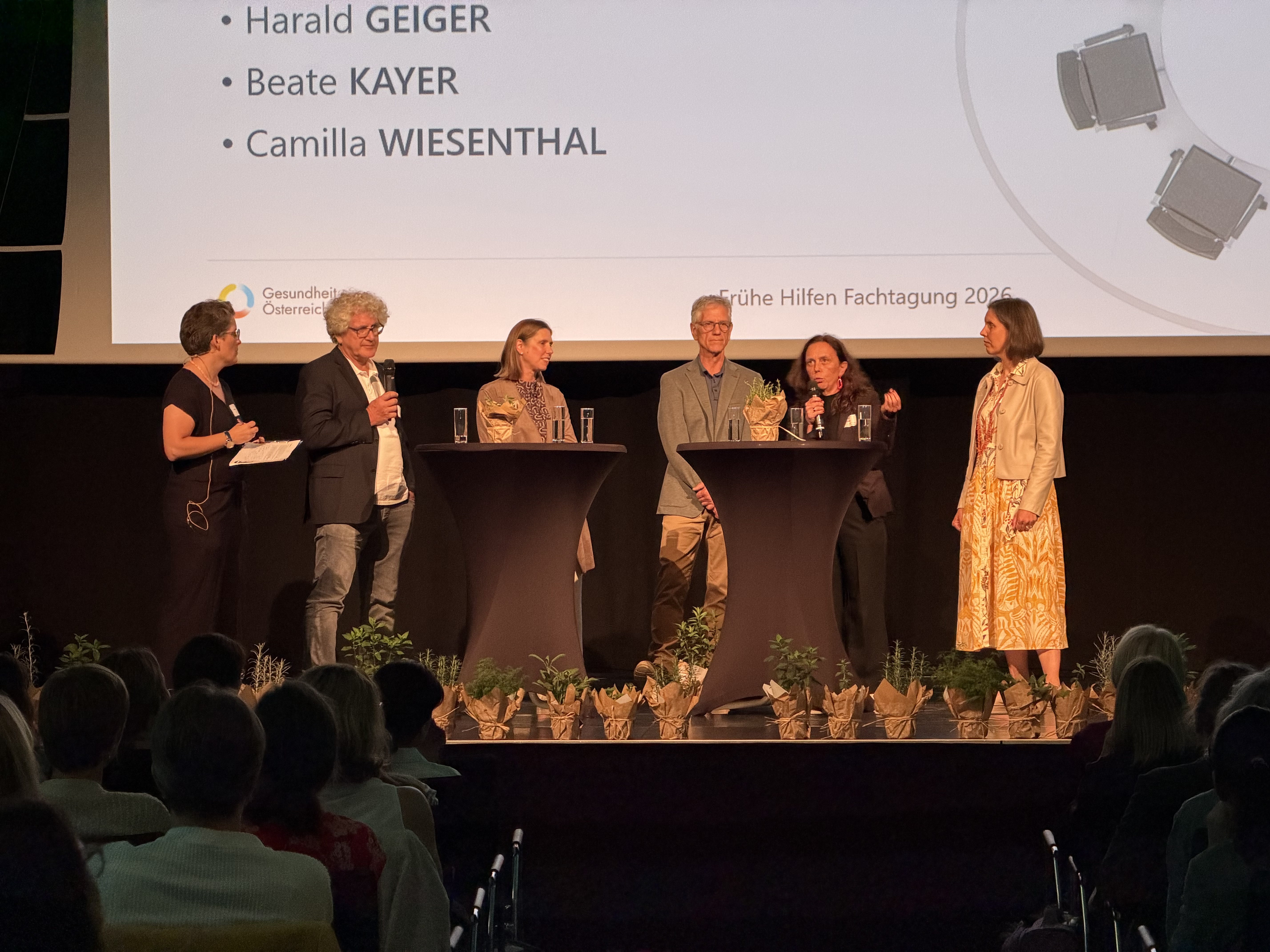 Podiumsdiskussion auf der Bühne der Fachtagung Frühe Hilfen 2026. Mehrere Personen stehen an Stehtischen und diskutieren vor Publikum. Im Hintergrund ist die Präsentationsfolie zur Diskussion sichtbar.