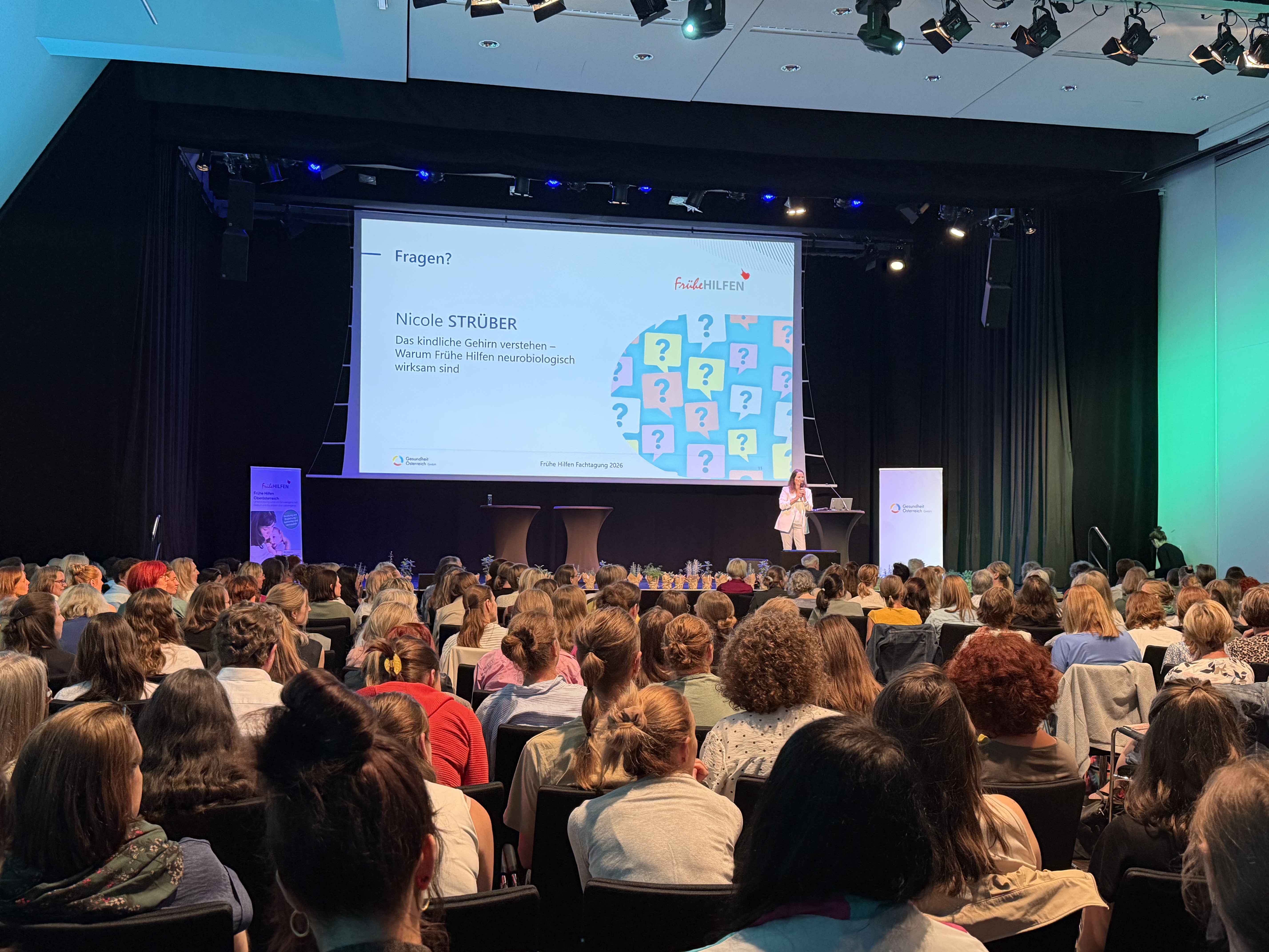 Vortrag im voll besetzten Veranstaltungssaal der Fachtagung Frühe Hilfen 2026. Eine Referentin präsentiert vor einer großen Leinwand mit dem Vortragstitel „Warum Frühe Hilfen neurobiologisch wirksam sind“. Die Zuhörenden verfolgen den Vortrag aus dem Publikum.