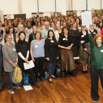 © NZFH.at Gruppenfoto von Teilnehmenden der österreichweiten Fachtagung Frühe Hilfen 2023. Zahlreiche Personen versammeln sich im Veranstaltungssaal; im Vordergrund wird die Resolution zur nachhaltigen Finanzierung der Frühen Hilfen symbolisch präsentiert.